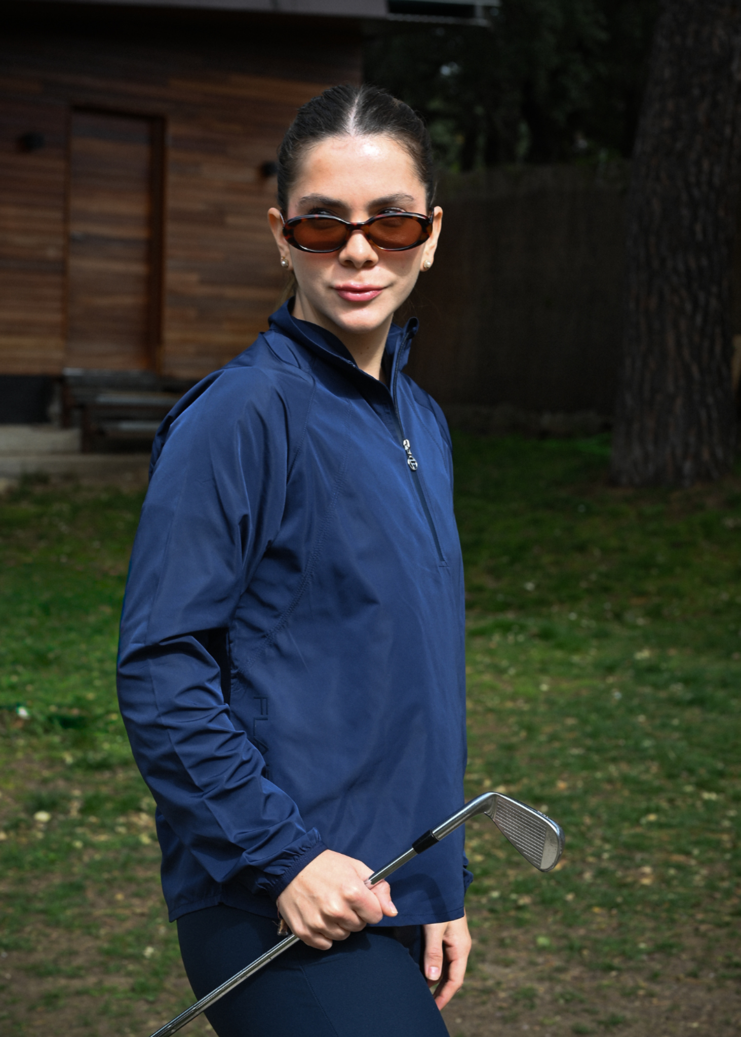 Person in a blue jacket holding a golf club outdoors with a wooden cabin in the background
