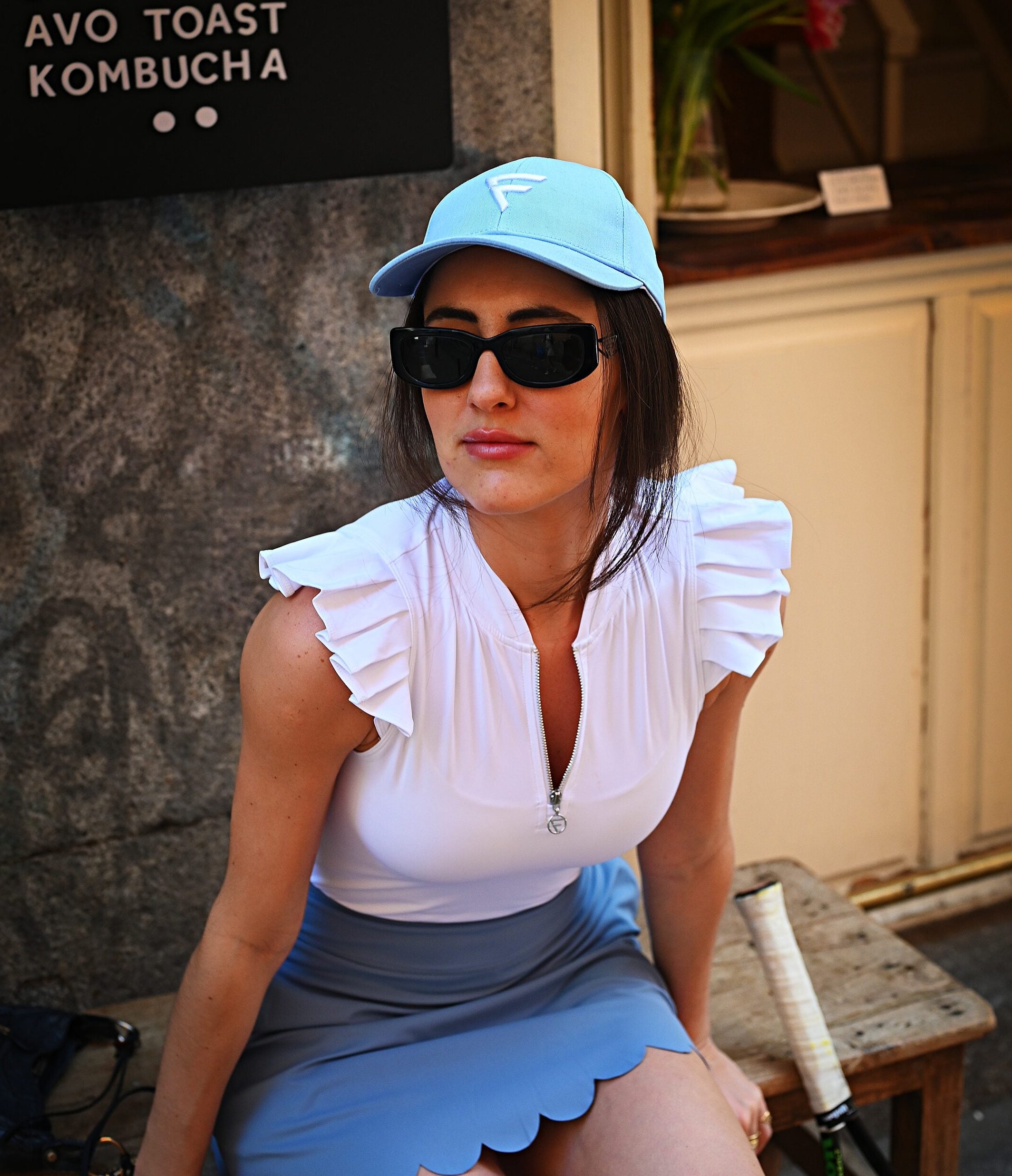 A woman wearing a light blue cap, black sunglasses, a white ruffled sleeveless top, and a light blue skirt sits on a bench in front of a bakery storefront. A black sign on the left lists bakery items including matcha, homemade chai, banana bread, cookies, granola, avocado toast, and kombucha.
