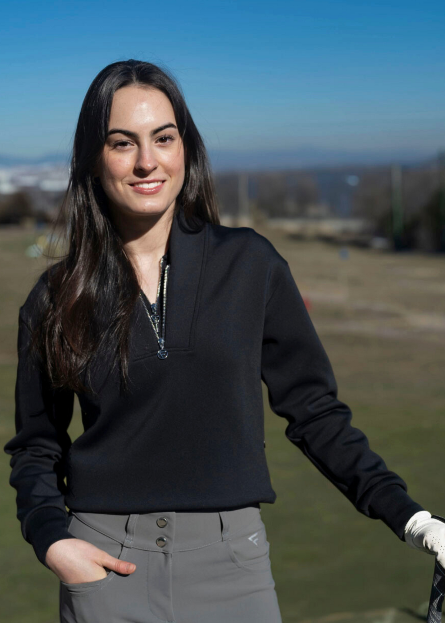 A woman golfer wearing a black sweater with a subtle logo on the side, standing on a golf course.