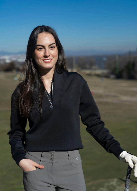 A woman golfer wearing a black sweater with a subtle logo on the side, standing on a golf course.